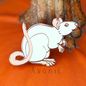 Op de afbeelding: Een witte emaille pin van een rat met een roségouden afwerking. De rat zit op zijn achterpoten met zijn staart om zijn lichaam gekruld. De pin is op een pompoen achtergrond.
