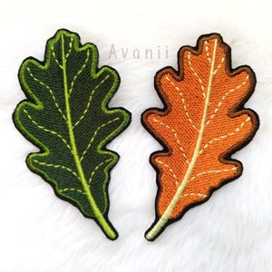 Pode incluir: Dois patches bordados em forma de folha de carvalho. Um é verde com costuras amarelas, o outro é laranja com costuras brancas. Ambos têm uma borda preta e estão sobre um fundo branco e fofo. A palavra "Avanii" está no canto superior direito.