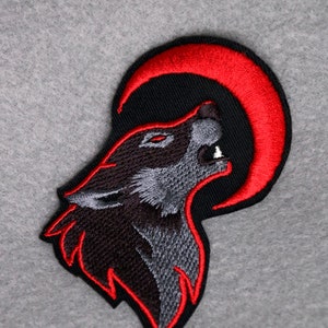 Op de afbeelding: Gestikt patch met een zwarte wolf die huilt naar een rode halve maan. De vacht van de wolf is gedetailleerd met grijze en zwarte stiksels. Het patch heeft een rode omtrek.