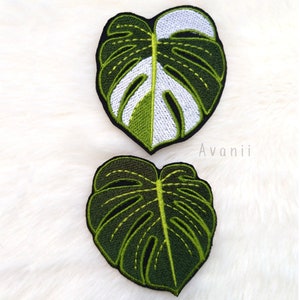 Può includere: Due toppe ricamate a forma di foglia di monstera. La toppa superiore è bianca e verde, mentre la toppa inferiore è completamente verde.