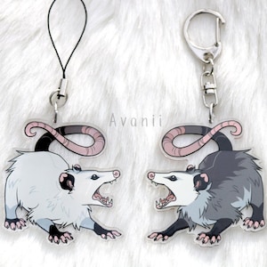 Op de afbeelding: Twee acryl opossum bedels met een zwart bandje en een zilveren sluiting. Eén opossum is lichtgrijs, de andere is donkergrijs, beide met roze staarten en open monden. De bedels zijn ontworpen in cartoonstijl.