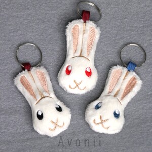 Op de afbeelding: Drie witte pluche konijnen sleutelhangers met geborduurde gezichten. Het konijn links heeft zwarte ogen, het konijn in het midden heeft rode ogen en het konijn rechts heeft blauwe ogen. Elk konijn heeft een ander gekleurd lint aan de sleutelring bevestigd.