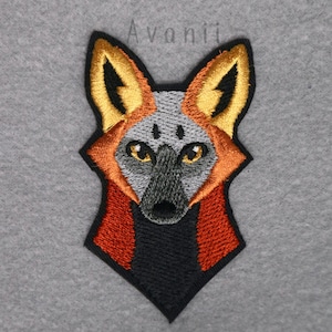 Pode incluir: Etiqueta bordada com a cabeça de um cão selvagem africano vermelho, preto e dourado. O cão tem o nariz preto e os olhos amarelos.