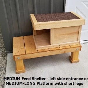 May include: A wooden bird feeder with a raised platform and a small shelter with a side entrance. The feeder is made of natural wood and has a brown roof.