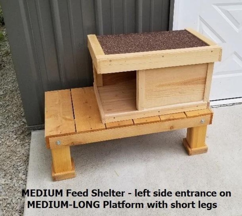May include: A wooden bird feeder with a raised platform and a small shelter with a side entrance. The feeder is made of natural wood and has a brown roof.