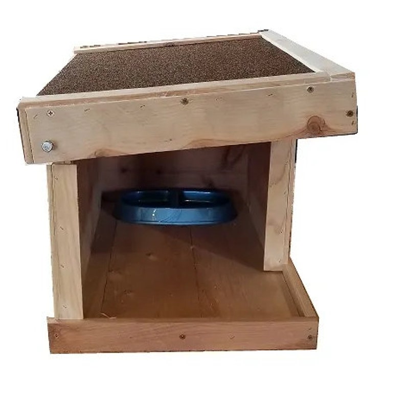May include: A wooden dog house with a slanted roof and a blue food bowl inside. The house is made of natural wood and has a simple design.