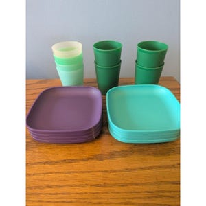 May include: A collection of colorful plastic dishware, including stacked plates in purple and teal, and cups in shades of green. The plates are square with rounded corners. The cups are stacked in groups of two and three.