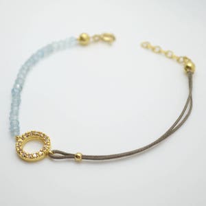 May include: A delicate bracelet with a gold circle charm set with clear stones. The bracelet is made with light blue beads and a brown cord.