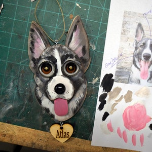 May include: A wooden ornament of a dog's face with black, white, and gray fur. The dog has brown eyes and a pink tongue sticking out. The ornament is hanging from a string and has a wooden heart with the word "Atlas" on it.