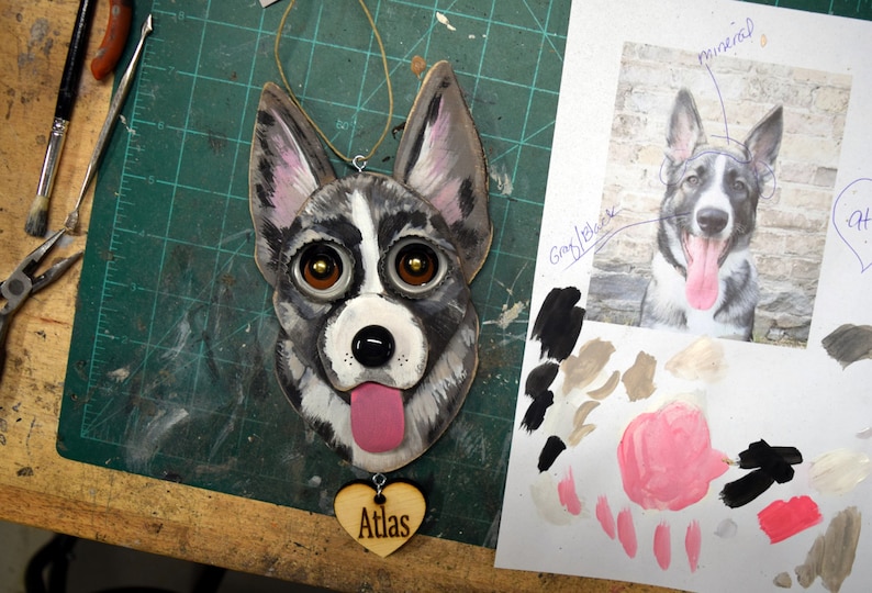 May include: A wooden ornament of a dog's face with black, white, and gray fur. The dog has brown eyes and a pink tongue sticking out. The ornament is hanging from a string and has a wooden heart with the word "Atlas" on it.