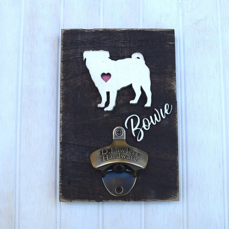May include: A wooden bottle opener with a white pug silhouette and the name "Bowie" in cursive. The bottle opener is made by D. Lawless Hardware.