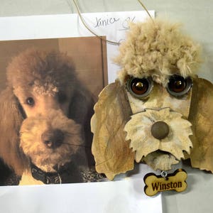 May include: A wooden ornament of a poodle dog with brown fur, brown eyes, and a brown nose. The ornament has a bone-shaped tag with the name "Winston" written on it.