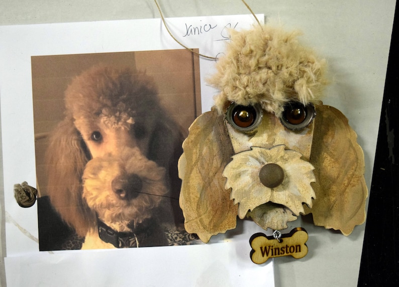 May include: A wooden ornament of a poodle dog with brown fur, brown eyes, and a brown nose. The ornament has a bone-shaped tag with the name "Winston" written on it.