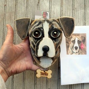 May include: A wooden dog head sculpture painted to resemble a brown and white dog with large brown eyes. The sculpture is attached to a wooden bone-shaped tag with the name "Stella" engraved on it.