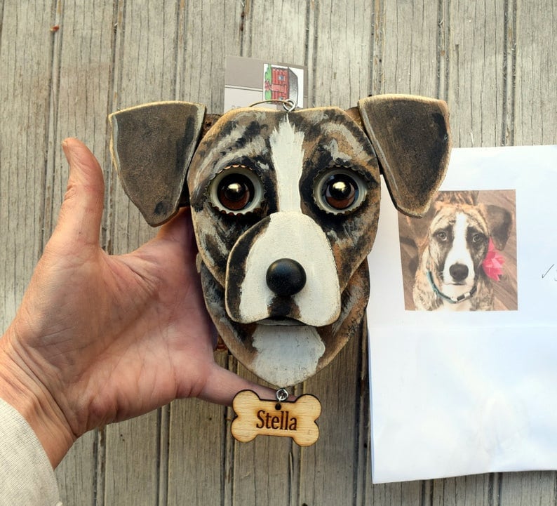 May include: A wooden dog head sculpture painted to resemble a brown and white dog with large brown eyes. The sculpture is attached to a wooden bone-shaped tag with the name "Stella" engraved on it.