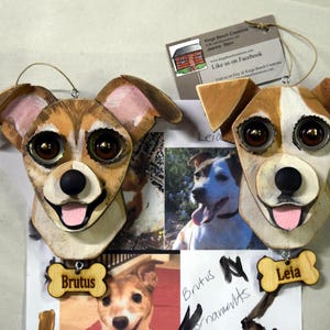 May include: Two wooden dog ornaments, one brown and white with brown eyes and a pink tongue, the other brown and white with brown eyes and a pink tongue. The ornaments are hanging from a string and have a wooden bone tag with the name "Brutus" and "Leia" written on them.