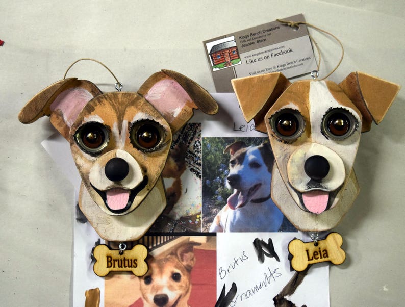 May include: Two wooden dog ornaments, one brown and white with brown eyes and a pink tongue, the other brown and white with brown eyes and a pink tongue. The ornaments are hanging from a string and have a wooden bone tag with the name "Brutus" and "Leia" written on them.