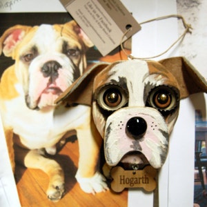 May include: A hand-painted wooden bulldog ornament with brown, white, and black paint. The ornament has large, expressive eyes and a pink nose. The ornament is hanging from a string and has a wooden tag that says "Hogarth".