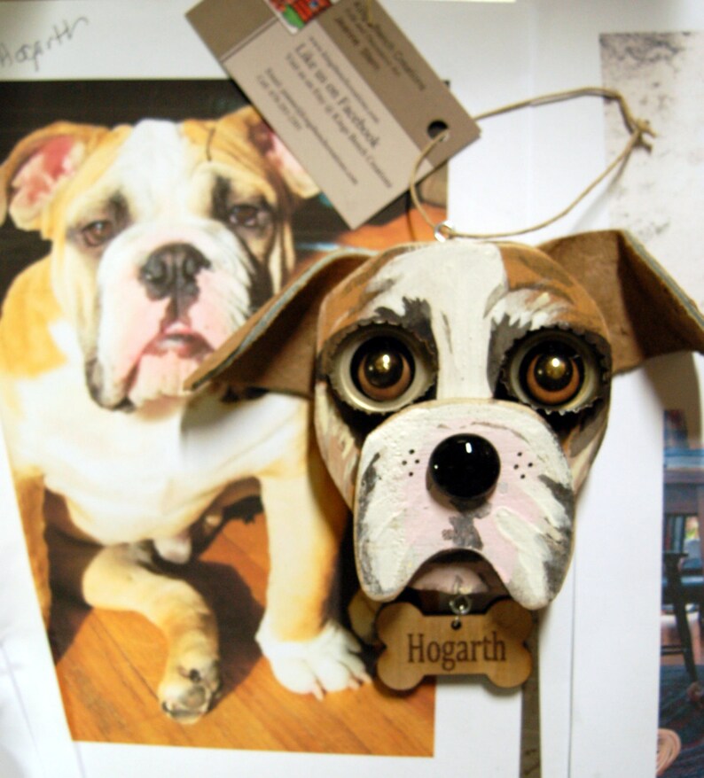 May include: A hand-painted wooden bulldog ornament with brown, white, and black paint. The ornament has large, expressive eyes and a pink nose. The ornament is hanging from a string and has a wooden tag that says "Hogarth".