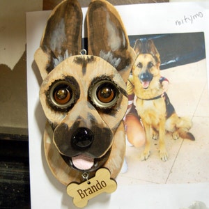 May include: A wooden German Shepherd dog head sculpture with bottle cap eyes and a pink tongue. The sculpture has a wooden bone tag that says "Brando".