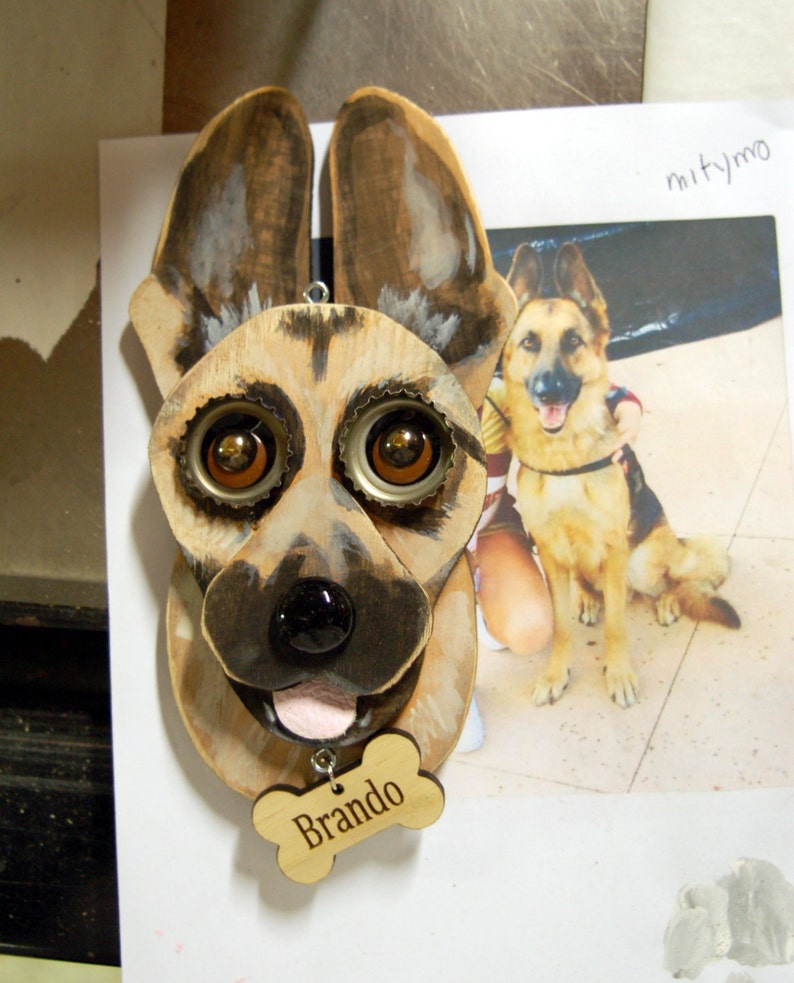 May include: A wooden German Shepherd dog head sculpture with bottle cap eyes and a pink tongue. The sculpture has a wooden bone tag that says "Brando".