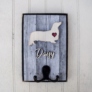 May include: A wooden dog leash hook with a gray and white woodgrain design. The hook is shaped like a dachshund with a red heart on its chest. The name "Daisy" is written in cursive on the hook.