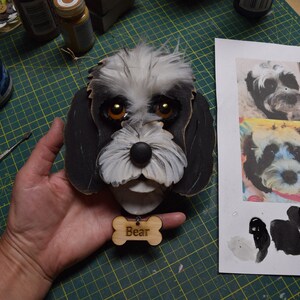 May include: A black and white wooden dog head with a name tag that says "Bear". The dog has brown eyes and a black nose. The dog is being held by a hand.