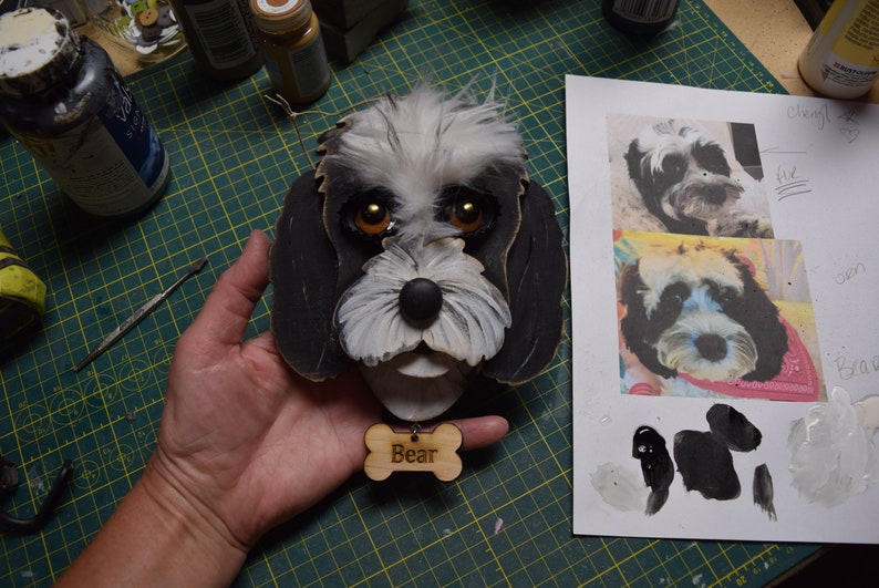 May include: A black and white wooden dog head with a name tag that says "Bear". The dog has brown eyes and a black nose. The dog is being held by a hand.