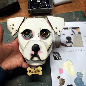 May include: White wooden dog head ornament with brown eyes, black nose, and pink painted details. The ornament has a wooden tag that says "Marigold".