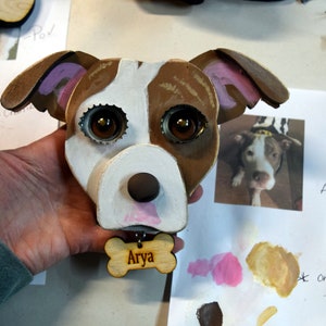 May include: A wooden dog head sculpture with brown, white, and pink paint. The dog has large brown eyes made from bottle caps and a pink nose. The sculpture has a wooden bone tag with the name "Arya" written on it.