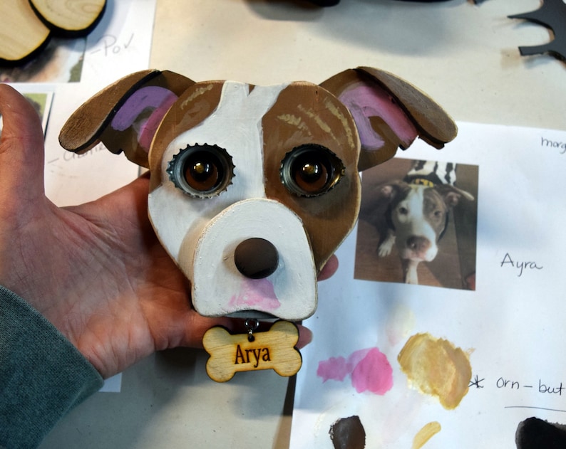 May include: A wooden dog head sculpture with brown, white, and pink paint. The dog has large brown eyes made from bottle caps and a pink nose. The sculpture has a wooden bone tag with the name "Arya" written on it.
