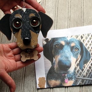 May include: A hand-painted wooden ornament of a dog's head with black and brown fur, large brown eyes, and a black nose. The ornament has a wooden bone charm with the name "Morgan" engraved on it.