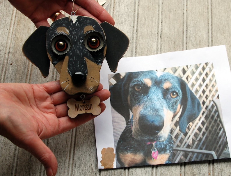 May include: A hand-painted wooden ornament of a dog's head with black and brown fur, large brown eyes, and a black nose. The ornament has a wooden bone charm with the name "Morgan" engraved on it.