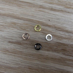 May include: Four metal rings in different finishes: gold, silver, rose gold, and black. The rings are arranged in a diamond shape on a wooden surface.