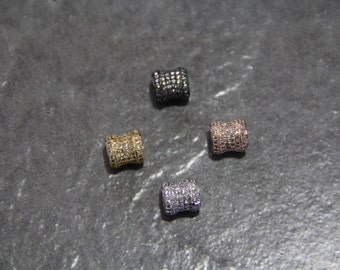 May include: Four small, cylindrical beads in gold, black, rose gold, and silver. Each bead is covered in small, sparkling stones. The beads are arranged on a dark gray surface, showcasing their varied colors and textures.