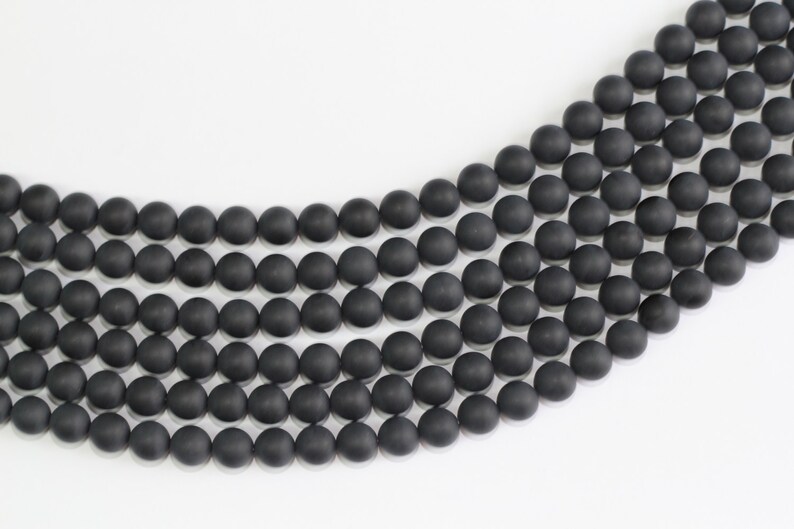 May include: A strand of black matte round beads, arranged in a curved pattern on a white surface.