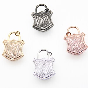 May include: Four lock charms with a shield shape, each with a different metal finish: black, gold, rose gold, and silver. Each charm is covered in clear rhinestones.