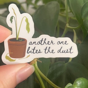 May include: A white sticker with a drawing of a wilted plant in a brown pot. The text "another one bites the dust" is written in black cursive.