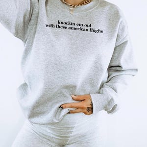 May include: A gray sweatshirt with the text "knockin em out with these american thighs" printed on the front. The sweatshirt is being worn by a person wearing gray shorts.