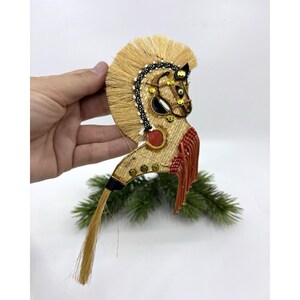 Puede incluir: Un adorno decorativo con forma de caballo hecho de fibras de color canela, con una crin de cuentas blancas y negras, lentejuelas doradas, un acento circular rojo y un largo fleco naranja. La pieza incluye una cola colgante de fibras color canela.