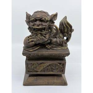 May include: A bronze-colored, detailed sculpture of a mythical creature, possibly a guardian lion, resting on a rectangular base. The creature has a fierce expression, holding a ball, with intricate carvings on the base.