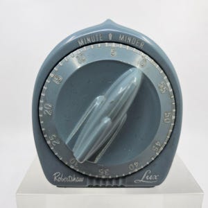 May include: A vintage blue Robertshaw Lux Minute Minder kitchen timer with a pointed top arch. The circular dial features white numbers from 0 to 55, with a central rotating knob. The text "MINUTE MINDER" is printed at the top, with brand names below.