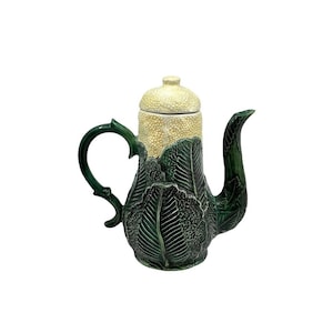 May include: A ceramic teapot with a leaf design. The teapot is primarily dark green with a textured leaf pattern. The lid and upper portion are a speckled cream color. The handle and spout are curved.