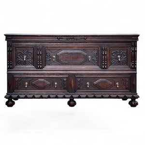 May include: A dark brown antique chest of drawers with ornate carvings and brass hardware. The chest features two drawers and decorative panels with floral and geometric designs. The chest rests on four rounded feet.