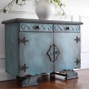 May include: A teal-colored antique cabinet with two drawers and two doors. The cabinet has ornate hardware and decorative accents. A white vase with flowers sits on top. The cabinet is placed against a white wall.