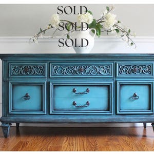 May include: A teal-colored wooden dresser with ornate carvings and dark hardware. The dresser has multiple drawers and cabinet doors. A white vase with white flowers sits on top. The word "SOLD" is written above the dresser.