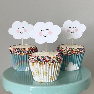 May include: Three cupcakes with white frosting and colorful sprinkles topped with cloud-shaped cake toppers with smiling faces. The cupcakes are on a light blue cake stand.