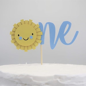 May include: A white cake with a blue "one" and a yellow sun with a smiling face and blue eyes. The sun is on a wooden stick.