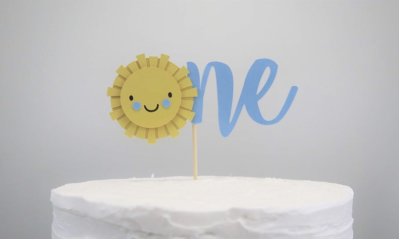 May include: A white cake with a blue "one" and a yellow sun with a smiling face and blue eyes. The sun is on a wooden stick.