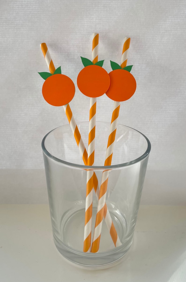 Peut inclure: Trois pailles en papier ray&eacute;es orange et blanc avec des d&eacute;coupes de fruits orange en haut. Les pailles sont dans un verre transparent.